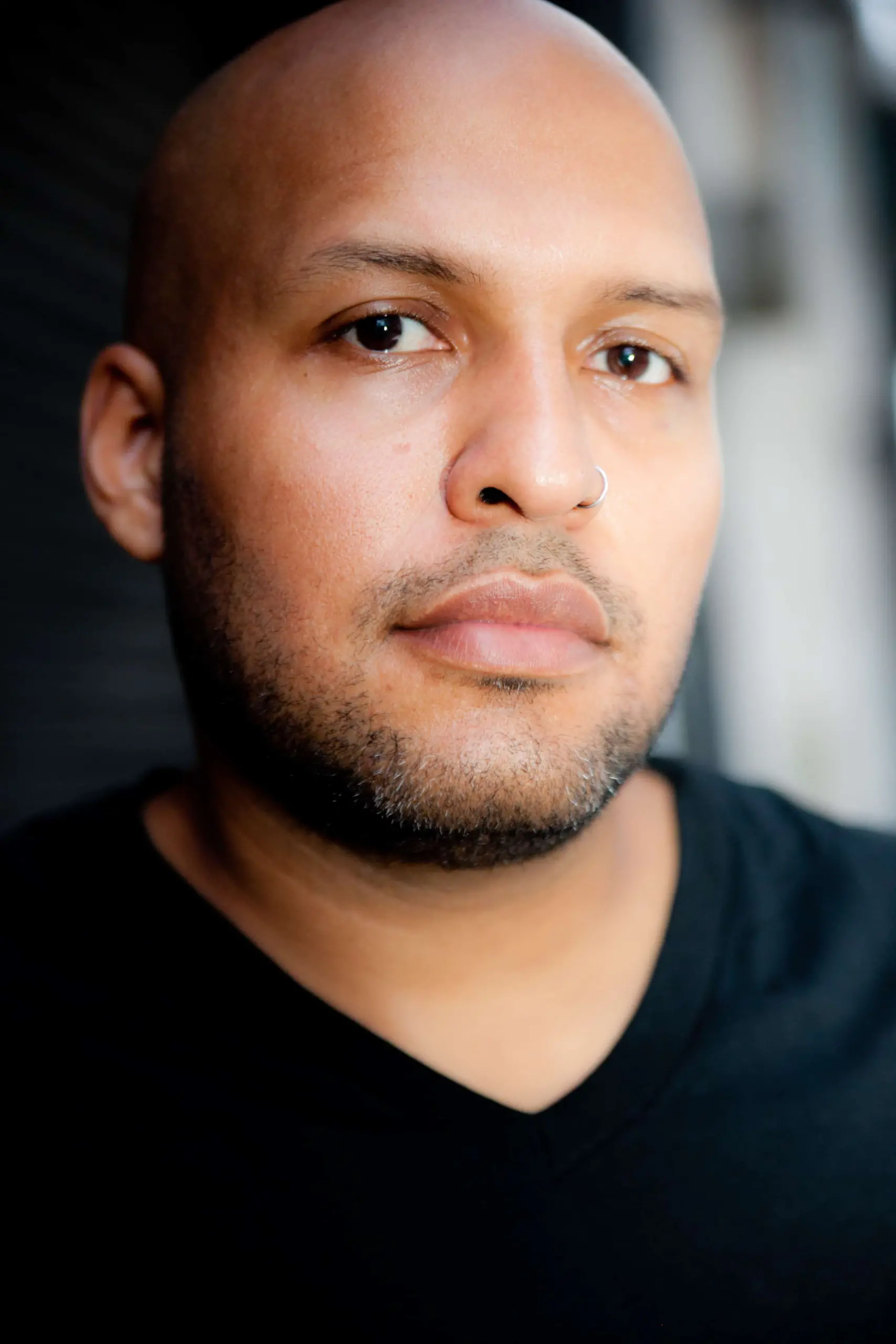 A person with a shaved head, short beard, and a nose ring, wearing a black V-neck shirt, looks calmly at the camera against a blurred background.