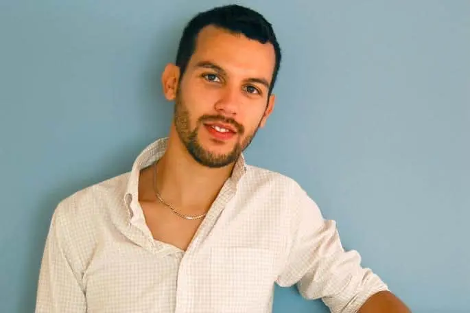 A young man with short dark hair and a beard stands against a light blue wall, wearing a light-colored, open-collared shirt and a chain necklace, looking at the camera with a slight smile.