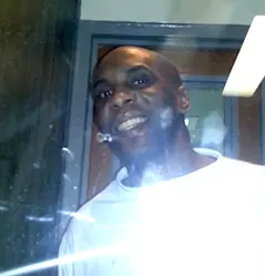 A man in a white shirt stands behind glass, smiling at the camera with a reflection and glare visible on the glass—a scene reminiscent of prison visits during covid. He is indoors, near a door and a dark wall.