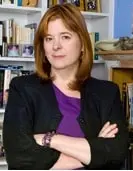 A woman with straight auburn hair, wearing a black blazer over a purple top, stands with her arms crossed in front of bookshelves filled with books and decorative items.