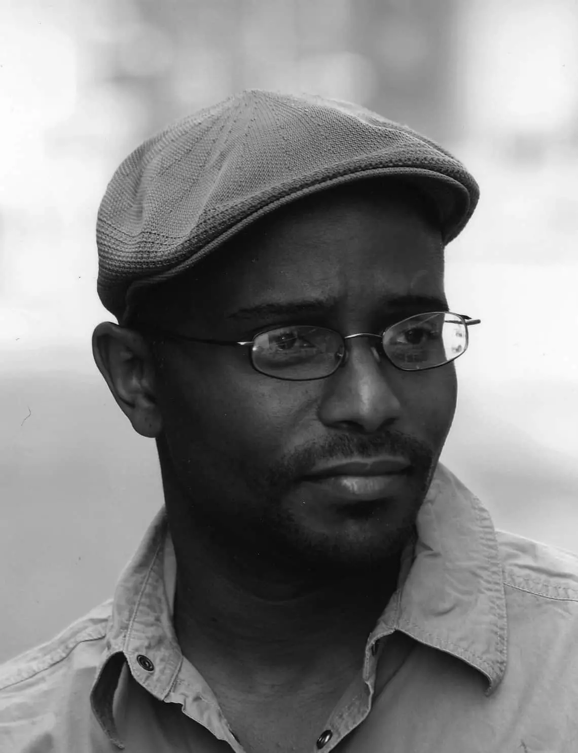 Black and white photo of a man wearing glasses and a flat cap, looking slightly to the side with a thoughtful expression. He is dressed in a collared shirt and has a trimmed beard. The background is blurred.
