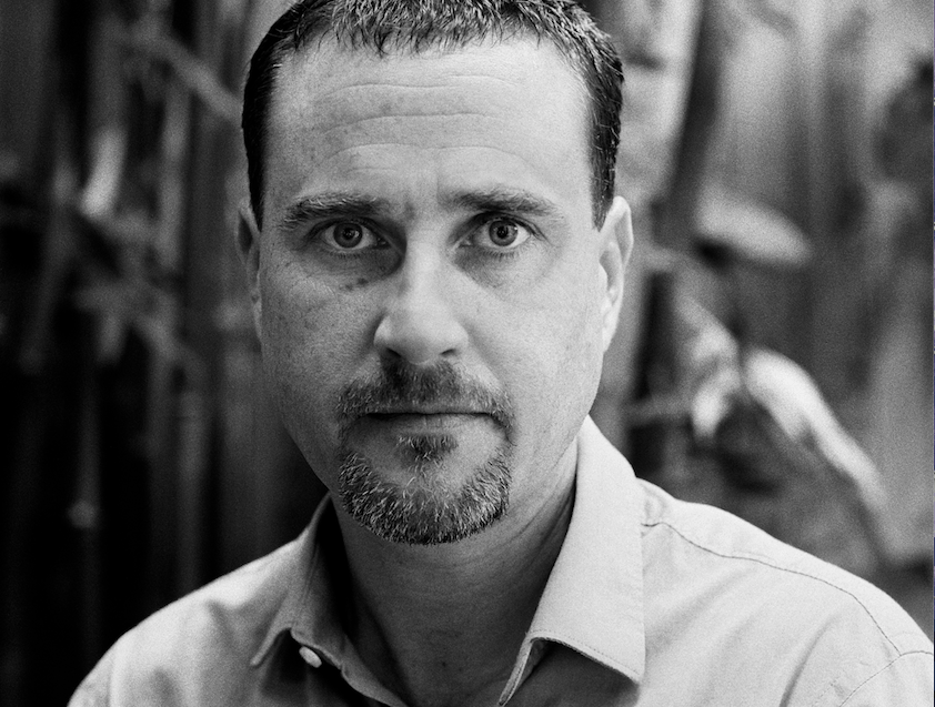 Black and white portrait of a man with short hair and a goatee, wearing a collared shirt, looking directly at the camera with a neutral expression. The background is blurred.