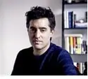 A man with dark hair and a dark sweater sits indoors in front of a bookshelf filled with various books. He is looking directly at the camera with a neutral expression.