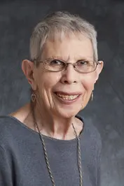 An older woman with short gray hair, glasses, and a gray top is smiling at the camera against a dark, neutral background. She is wearing earrings and a long necklace.