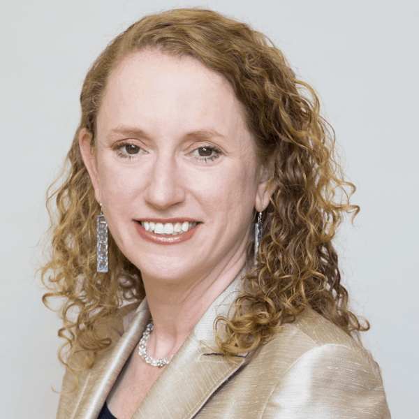 A woman with curly light brown hair smiles at the camera, wearing a beige blazer, long silver earrings, and a silver necklace—reflecting the spirit of NYC Free Expression Advocacy Institute PEN against a plain light background.