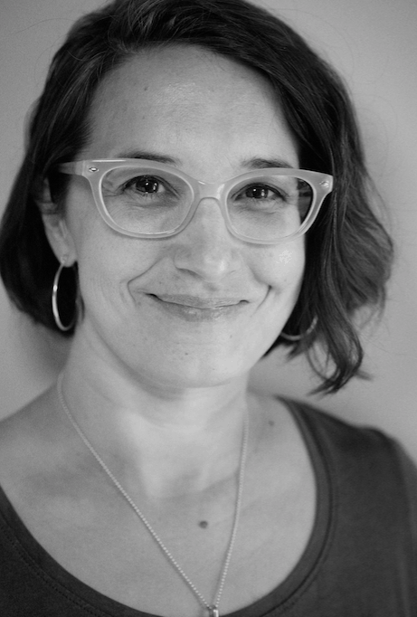 A person with short, dark hair and glasses smiles softly. They wear hoop earrings, a necklace, and a crew neck top. The portrait is black and white.