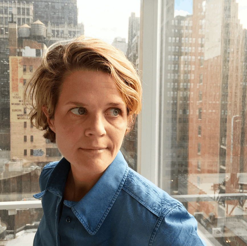 A person with short, light brown hair wearing a blue button-up shirt looks to the side while standing by a window with a cityscape of tall buildings in the background.