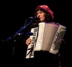 A person playing an accordion and singing into a microphone on a dark stage, illuminated by stage lights.