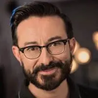 A man with dark hair, a neatly groomed beard, and glasses is smiling slightly. He is wearing a dark shirt, and the blurred background features warm, circular lights.