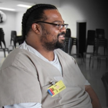A man with braided hair, glasses, and a beard sits smiling in a room with stacked chairs. He wears beige scrubs and a name tag with colored cards attached.