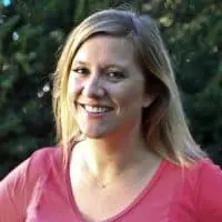 A woman with long blonde hair smiles at the camera. She is wearing a pink top and standing outdoors, with green foliage in the blurred background.