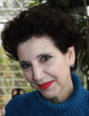 A woman with short curly dark hair, wearing a blue turtleneck sweater and red lipstick, smiles at the camera. The background shows large windows with natural light and green plants.