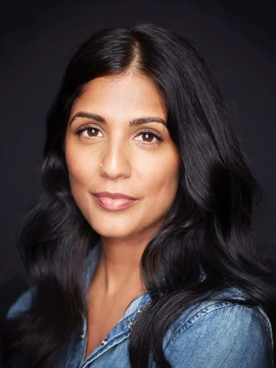 A woman with long, dark wavy hair and medium skin tone looks at the camera with a calm expression. She is wearing a denim shirt and has a small nose ring. The background is dark and plain.