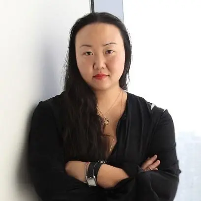 A woman with long dark hair and red lipstick stands with arms crossed against a white wall, wearing a black top and a watch on her left wrist. She has a neutral expression.