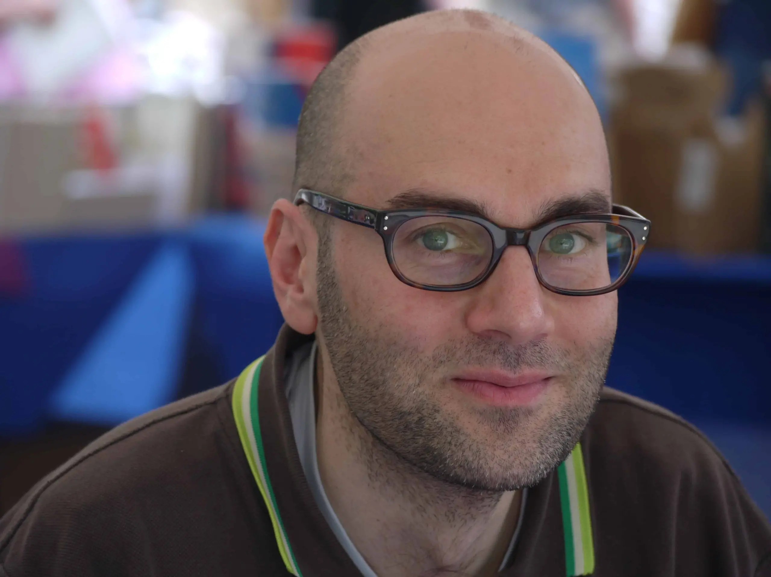 A man with glasses and a receding hairline is wearing a dark brown shirt with green trim. He is looking at the camera and smiling gently, with a blurred background of indoor objects behind him.