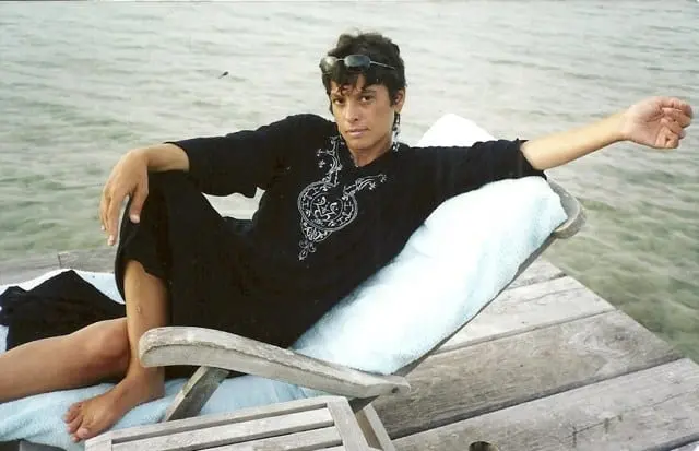 A person in a black embroidered outfit and snorkel mask relaxes on a lounge chair with a blue towel, on a wooden deck by clear, shallow water.