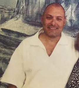 A smiling man with a shaved head wearing a white collared shirt stands in front of a painted backdrop. Only part of another person’s arm is visible on the right side of the image.