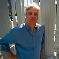 A man with gray hair wearing a blue button-up shirt stands outdoors, resting his arm on a white pillar, with more pillars and sunlight visible in the background.