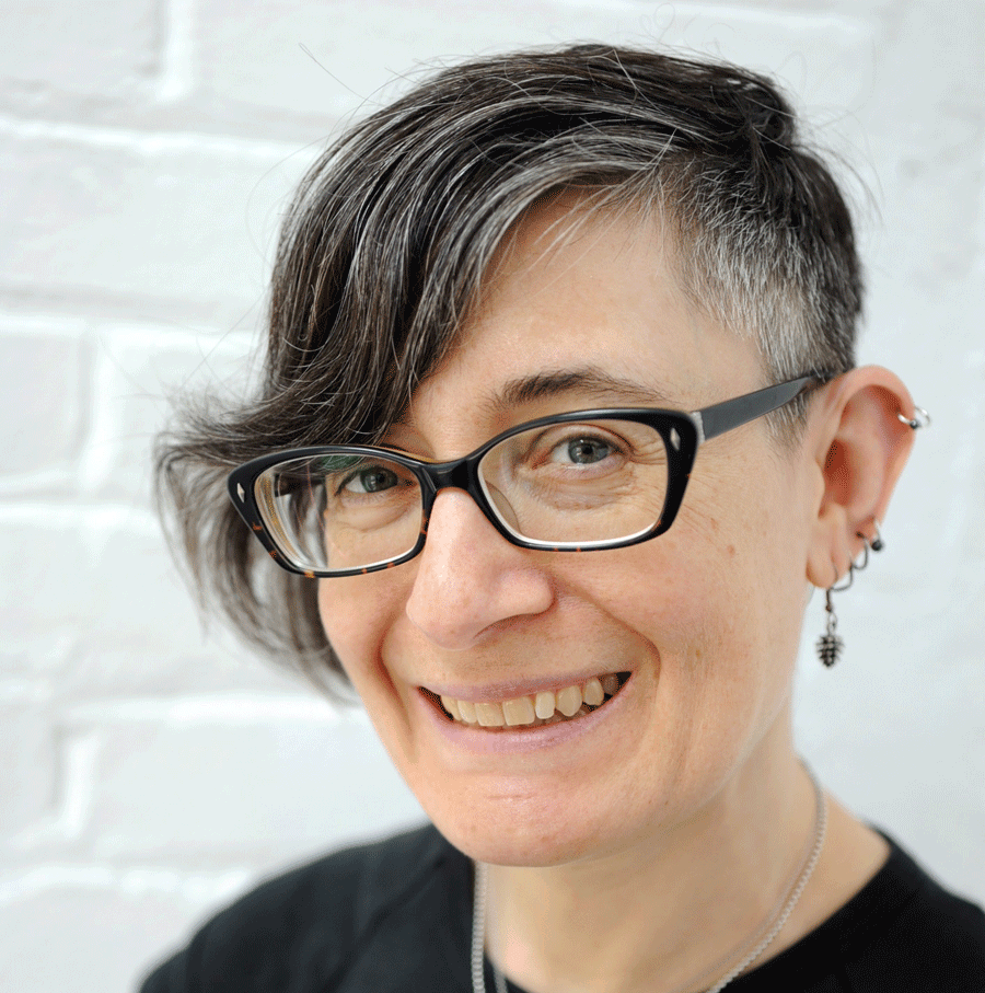 A person with short, gray and dark hair, wearing black-rimmed glasses, earrings, and a black shirt, smiles at the camera in front of a white brick wall.