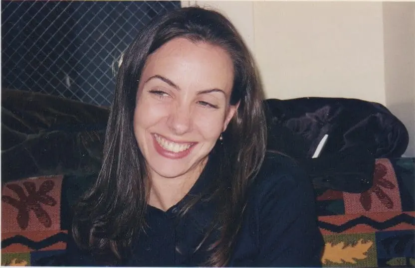 A woman with long brown hair and a black top is smiling and looking slightly to the side. She is sitting on a patterned sofa with a fence visible in the background.