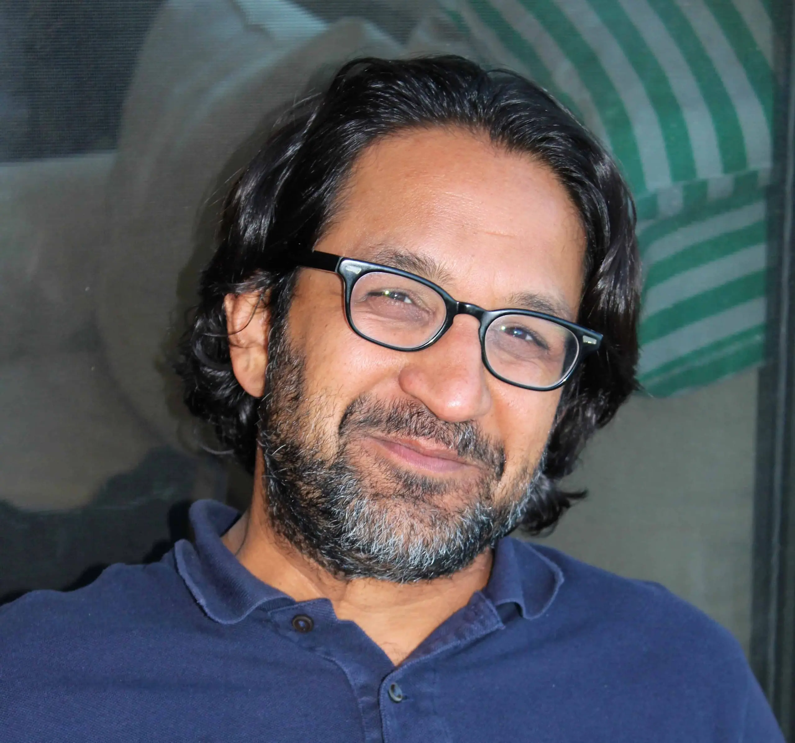 A man with dark hair, glasses, and a beard smiles at the camera. He is wearing a navy blue polo shirt, and the background includes a blurry green-and-white striped object.