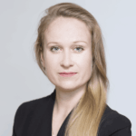 Portrait of a woman with long blonde hair, wearing a black blazer, looking directly at the camera with a neutral expression, evoking the theme of Discourse in Danger against a plain light background.