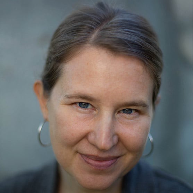 A woman with light skin, light brown hair pulled back, and blue eyes smiles softly at the camera. She is wearing hoop earrings and a grey top, with a neutral, blurred background.