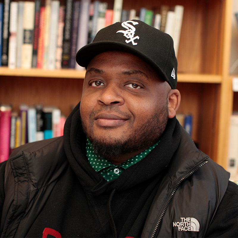 A man wearing a black jacket, black hoodie, and black White Sox baseball cap sits in front of a bookshelf, smiling slightly.