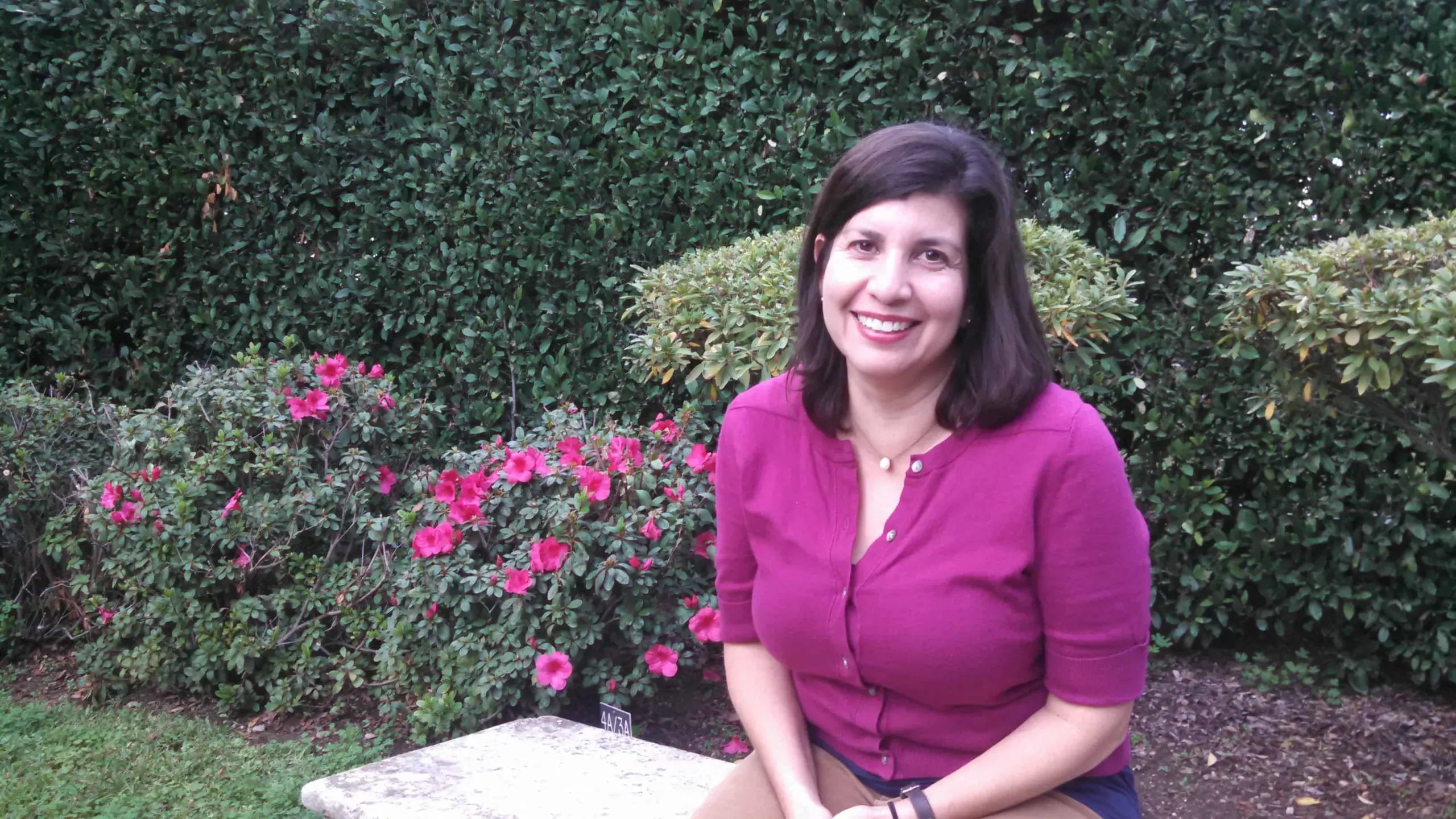 A woman with shoulder-length dark hair, wearing a magenta blouse, sits on a stone bench in a garden with flowering pink bushes and green shrubs, smiling at the camera.