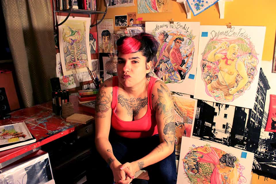 A tattooed person with pink and black hair sits on a stool in an art-filled studio, surrounded by colorful, detailed illustrations and drawing supplies, with a focused, contemplative expression.