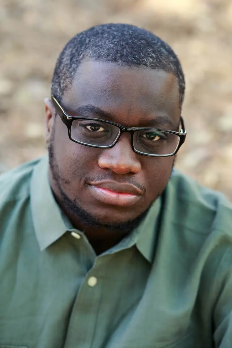 A man with short hair, glasses, and a short beard looks at the camera with a neutral expression. He is wearing a green collared shirt and is outdoors with a blurred background.