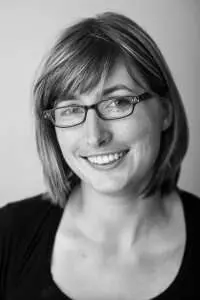 A woman with straight, shoulder-length hair, wearing glasses and a dark top, smiles while looking at the camera. The image is in black and white.