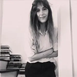 A woman with long hair, wearing a light-colored shirt and dark pants, stands indoors with her arms crossed and smiles. Stacks of books are visible in the background. The photo is in black and white.