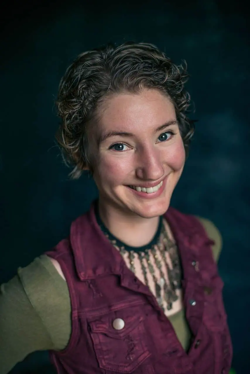 A person with short curly hair smiles at the camera, wearing a maroon vest over a green shirt and a beaded necklace, set against a dark, softly blurred background.