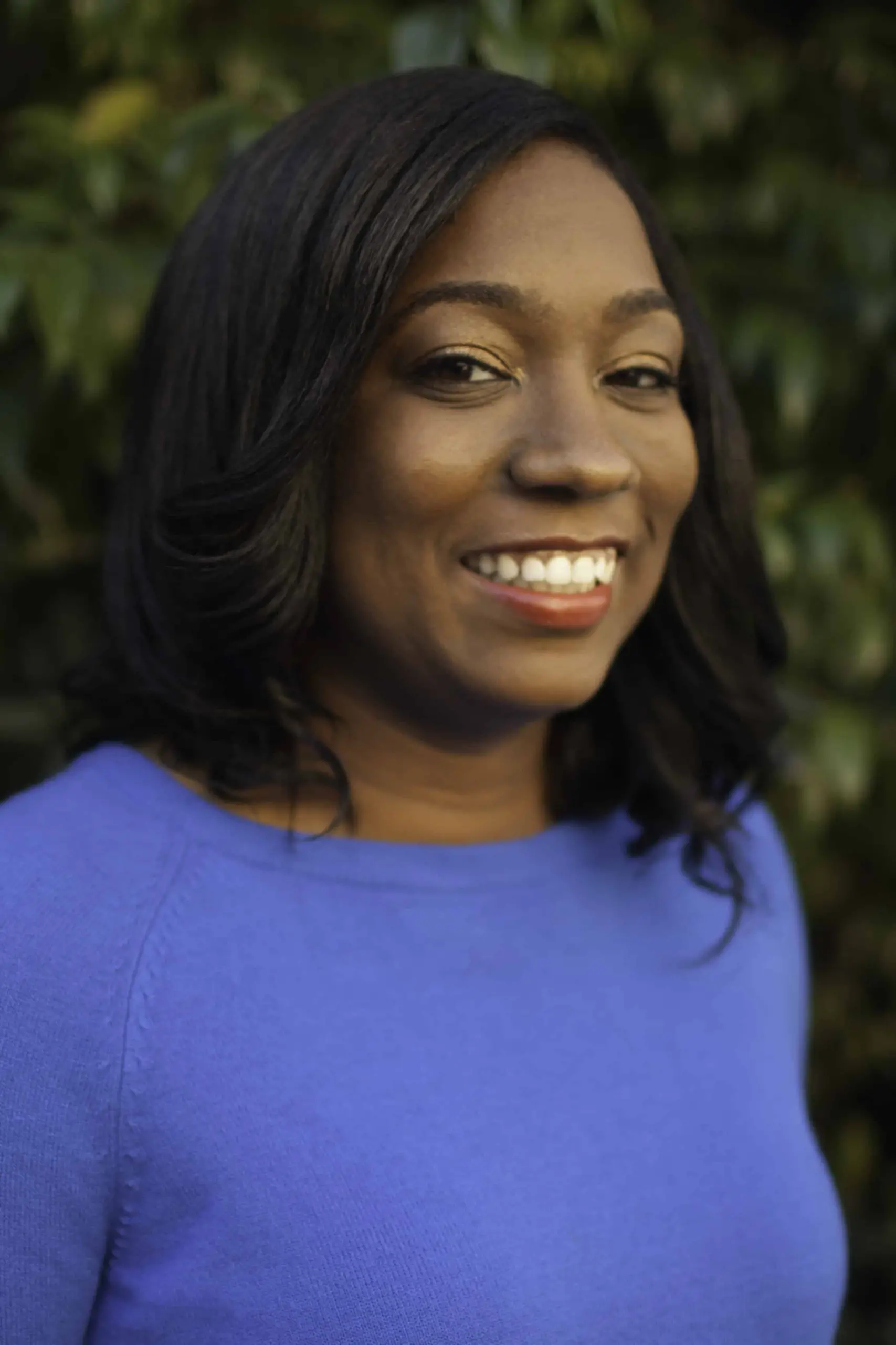A woman with shoulder-length black hair and a bright smile is wearing a blue top. She stands outdoors in front of green foliage, looking slightly to the side.