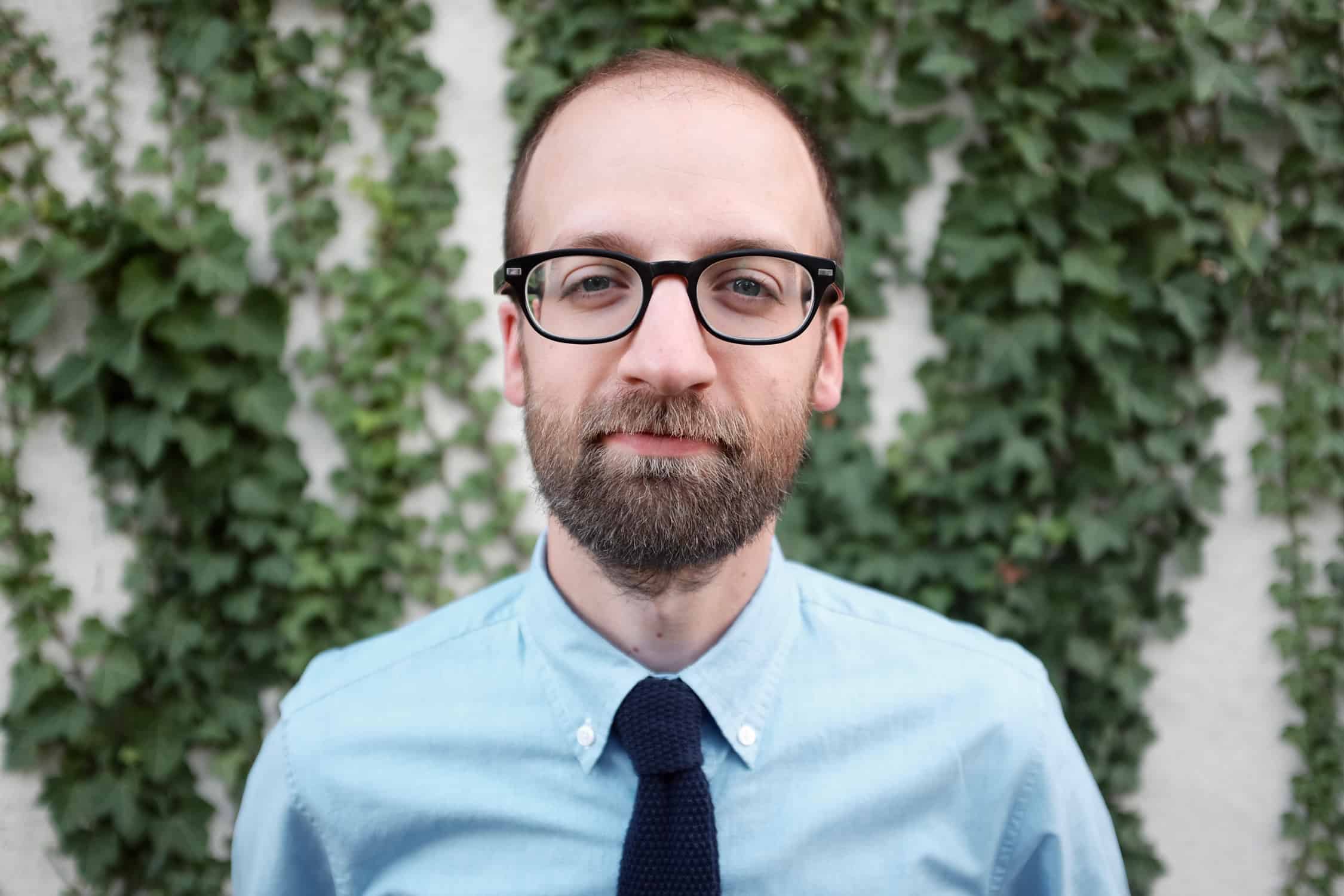 A man with a beard and glasses, wearing a light blue shirt and dark tie, stands in front of a wall covered with green ivy.