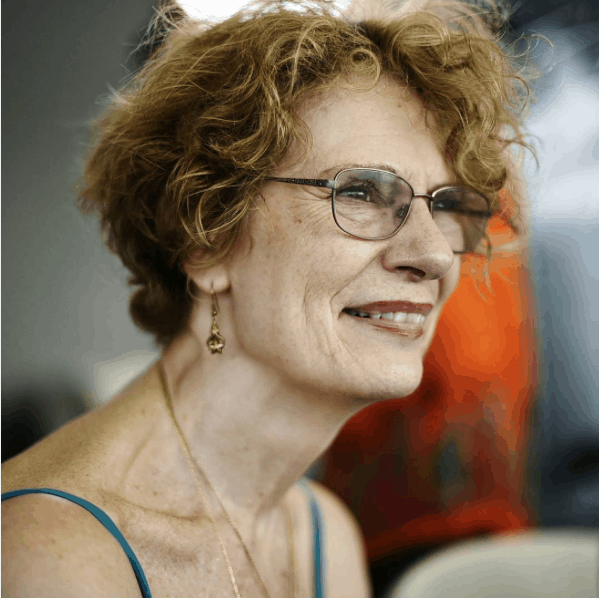 A person with short, curly light brown hair, glasses, and gold earrings smiles while looking to the side. They are wearing a thin-strapped top and a necklace. The background is blurred with a hint of orange.