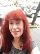 A person with long red hair smiles outdoors on a city street, with parked cars and trees visible in the background.