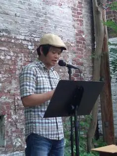 A person wearing a light-colored cap and plaid shirt reads or speaks into a microphone, standing at a podium outdoors in front of a weathered brick wall and trees.