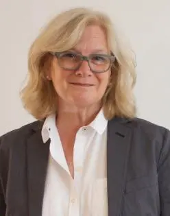 A woman with shoulder-length blonde hair and glasses is wearing a white shirt and a dark blazer, standing against a plain light background and smiling slightly.