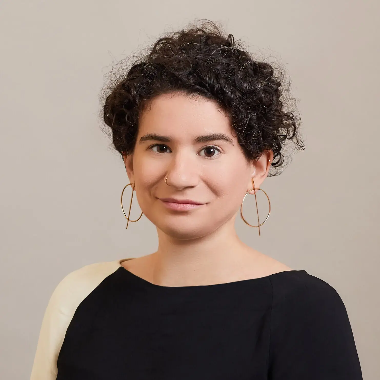 A person with short, curly dark hair poses in front of a plain beige background, wearing large hoop earrings and a black top with a light-colored accent on one shoulder.