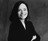 A woman with shoulder-length dark hair smiles at the camera, wearing a dark blazer and standing with her arms crossed in front of a plain, dark background.