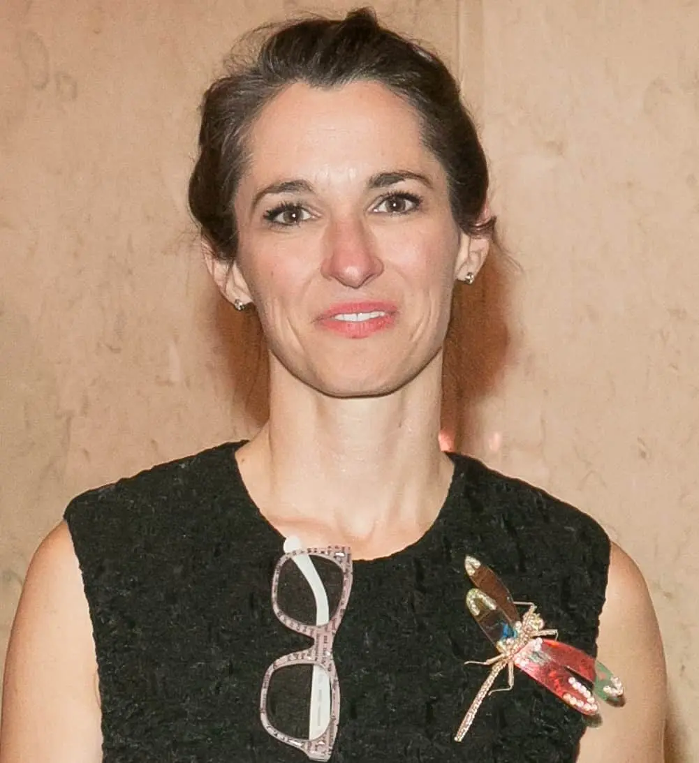 A woman with dark hair pulled back is wearing a sleeveless black textured top, glasses hanging from her collar, and a large decorative dragonfly brooch. She stands in front of a beige, slightly blurred background.