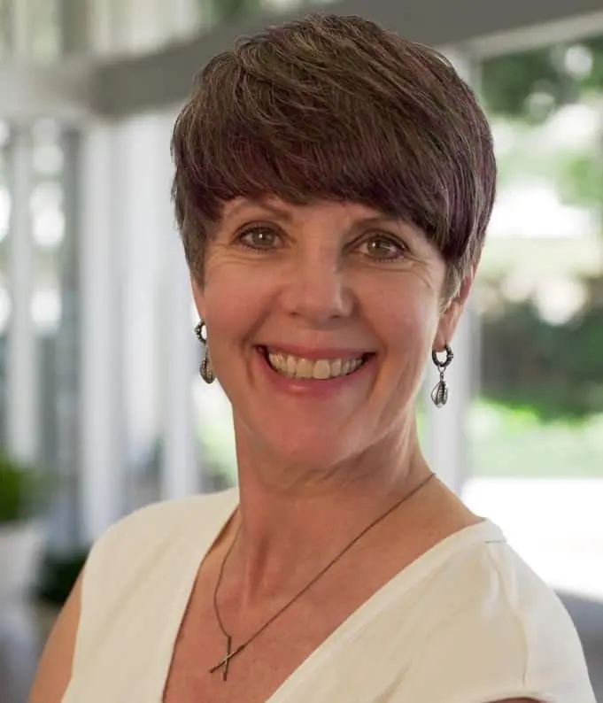 Smiling woman with short brown hair, wearing a white top, drop earrings, and a necklace with a cross pendant, standing indoors with blurred windows and greenery in the background.