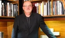 A man wearing glasses and a dark suit stands in front of wooden bookshelves filled with books, smiling at the camera.