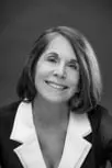 Black and white portrait of a woman with shoulder-length hair, smiling gently and wearing a dark blazer with a light-colored collar, set against a plain background.
