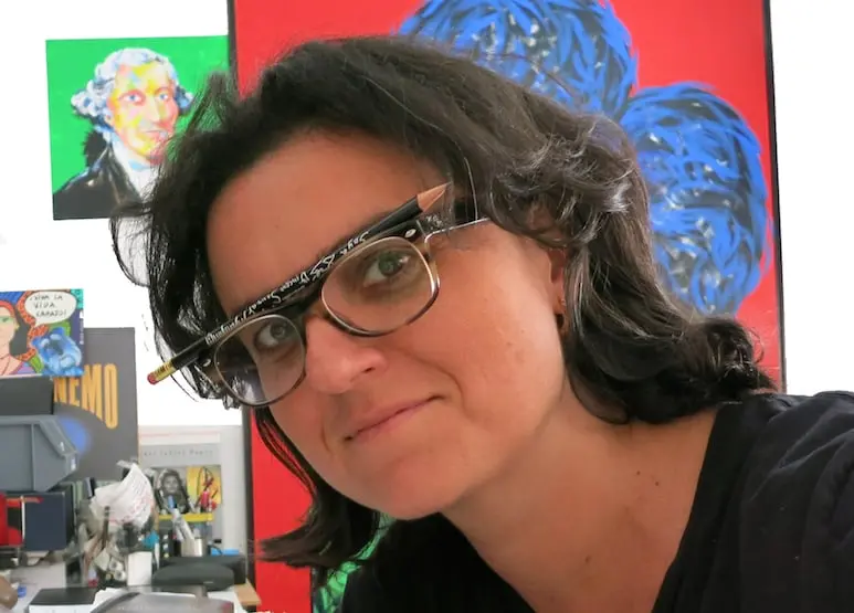 A woman with dark hair and glasses has a pencil balanced between the frames and her face. Colorful paintings and a cluttered desk are visible in the background.