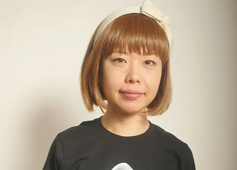A woman with straight, light brown hair in a bob cut wears a black shirt and a large cream-colored headband with a bow, standing in front of a plain, light background.