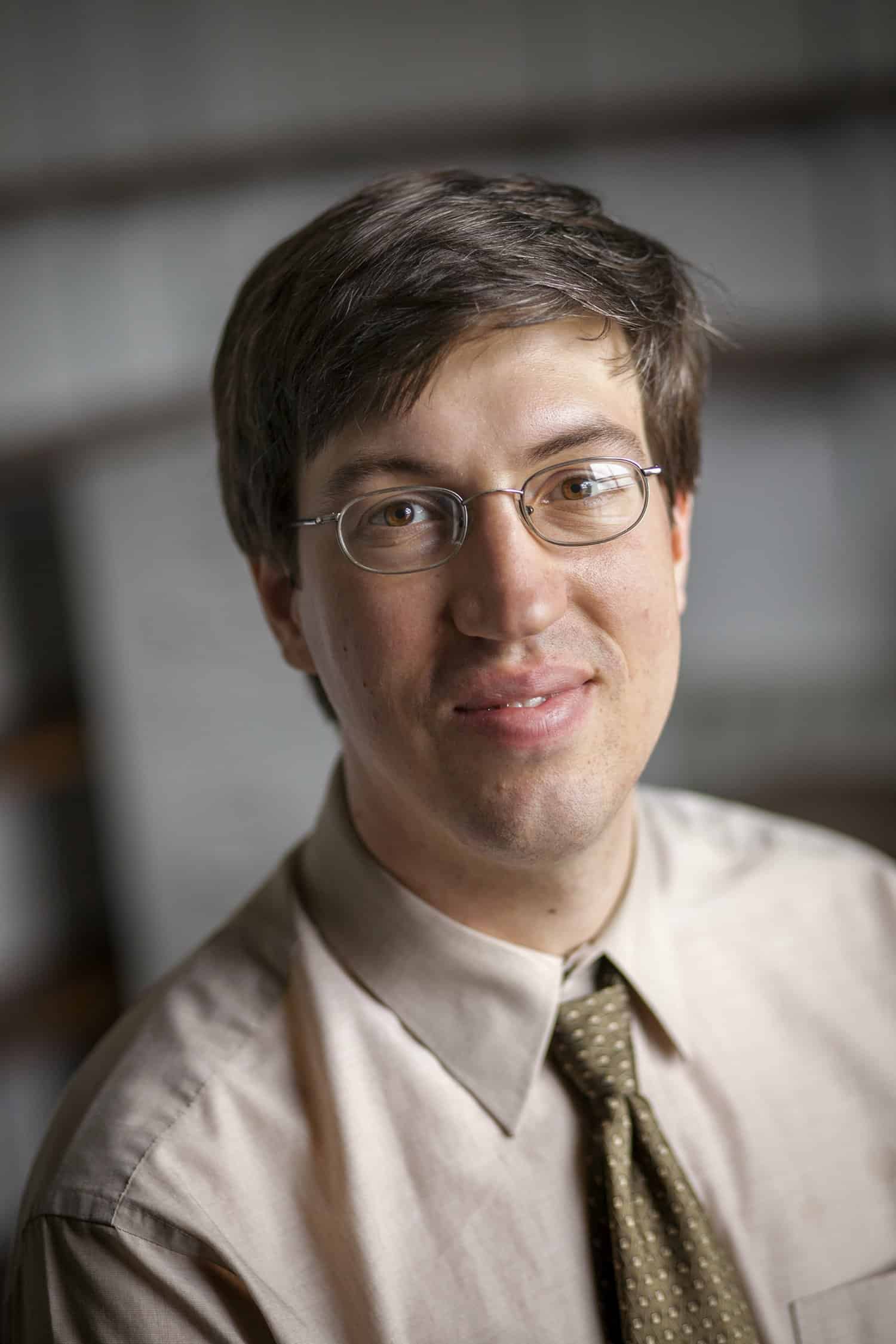 A man with short brown hair and glasses, wearing a beige shirt and patterned tie, smiles slightly while looking at the camera. The background is softly blurred.