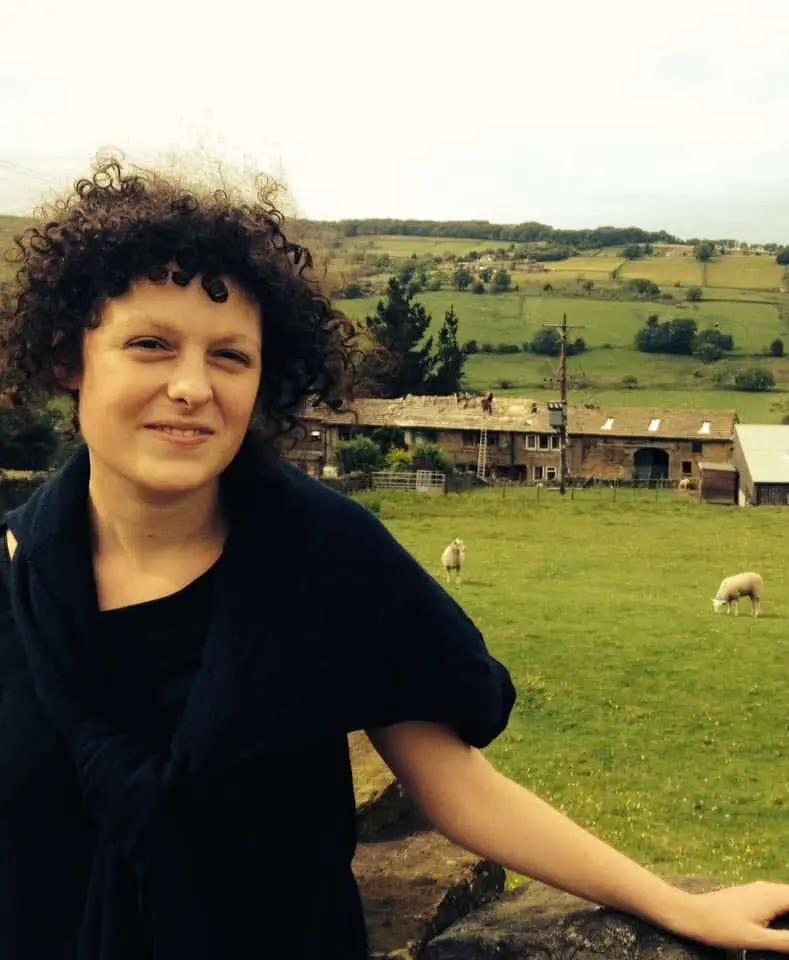 A woman with curly hair, wearing a black top and scarf, stands in front of a stone wall in a green field with sheep. Farm buildings and rolling hills are visible in the background under a cloudy sky.